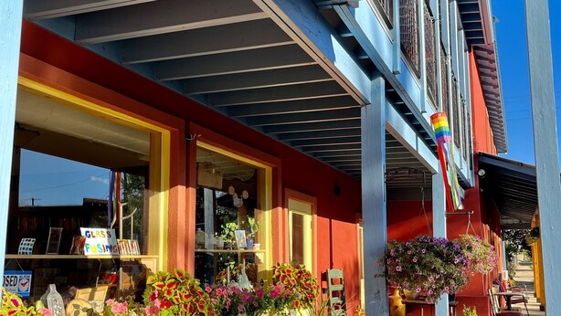 Un edificio con fachada roja y amarilla, un área de estar al aire libre cubierta con una mesa y sillas, y macetas de flores colgantes.