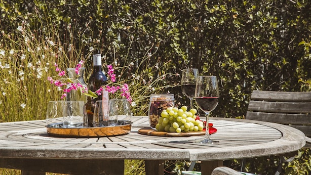 A round wooden table set with a bottle, glasses, a jar of grapes, and a glass of red wine, surrounded by greenery. - Mataro Cottage (Tanunda)