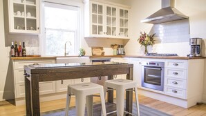 A modern kitchen with white cabinetry, a wooden island, two white bar stools, a stainless steel range hood, and a built-in oven. - Mataro Cottage (Tanunda)