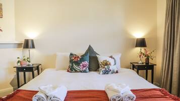 Een netjes opgemaakt bed met bloemenkussens, een rode plaid en opgerolde handdoeken. Twee nachtkastjes met lampen en een bloevas.