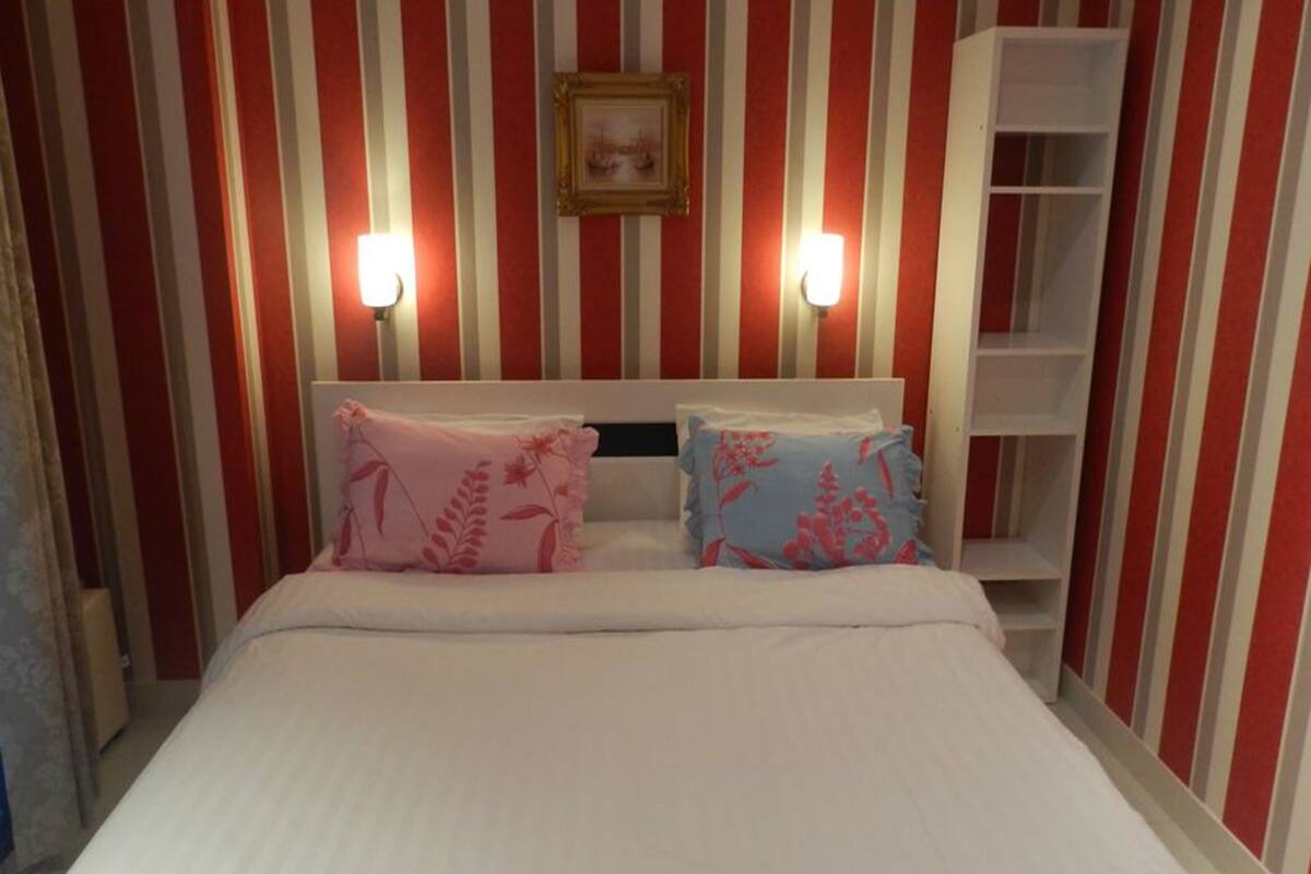 Una habitación de hotel con pared a rayas rojas y blancas, una cama con ropa blanca, dos almohadas con diseños florales y una mesita de noche blanca.