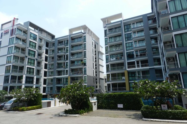 A multi-story residential building with balconies, a parking area, and greenery.