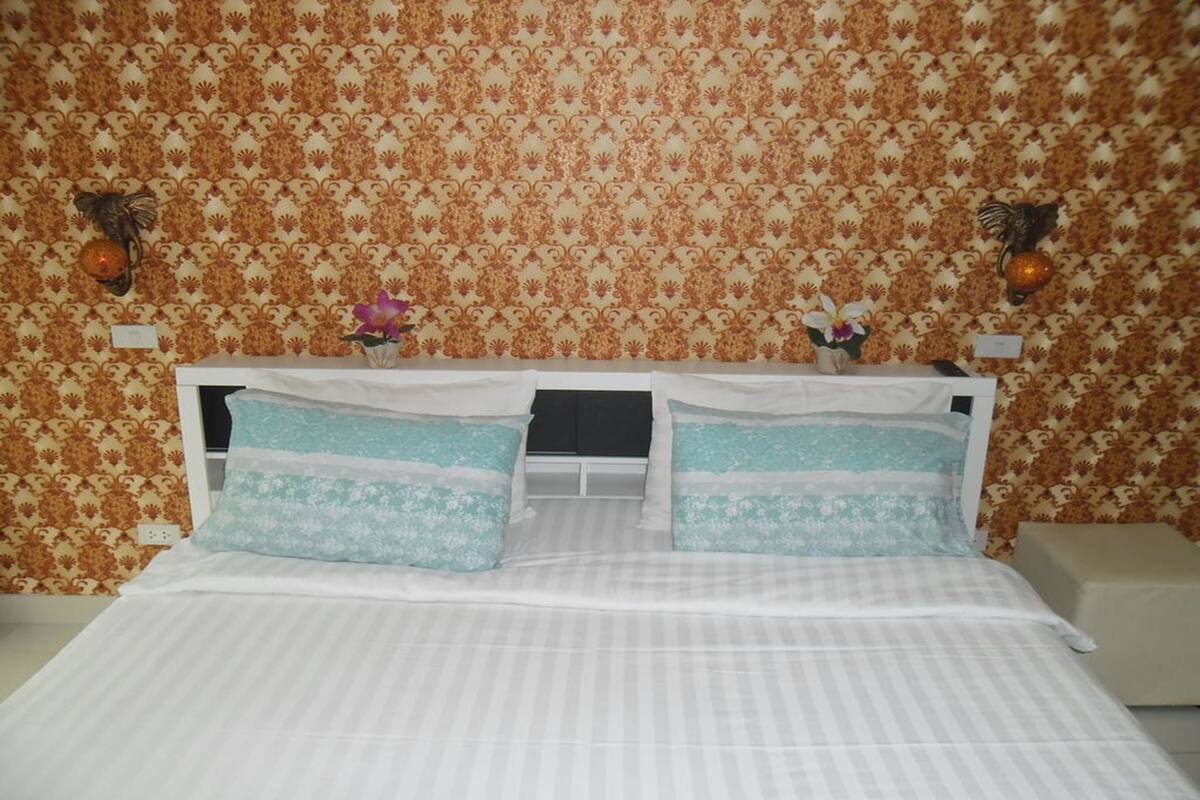 Una cama bien hecha con ropa de cama a rayas blanca y azul claro, dos jarrones decorativos con flores, y un fondo de papel tapiz estampado.