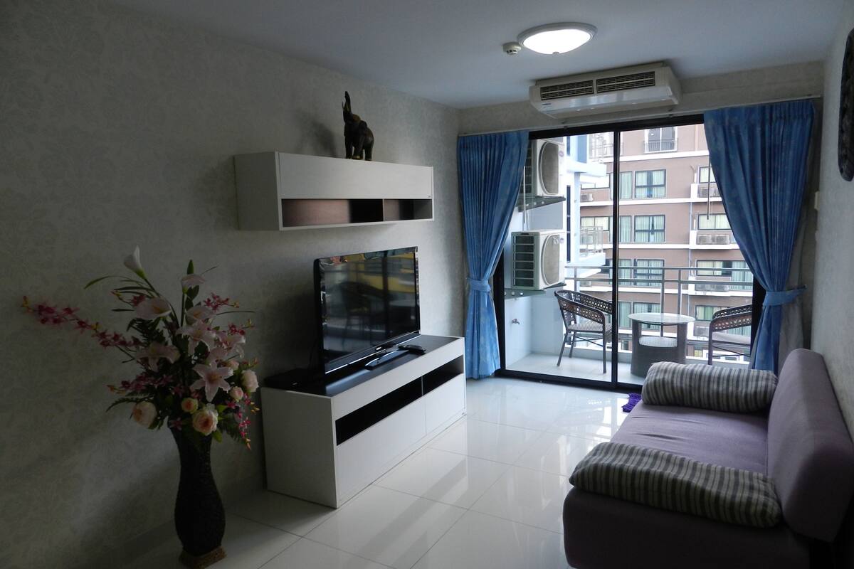Una sala de estar moderna con una televisión de pantalla plana, un sofá, un jarrón decorativo y un balcón con vistas a la ciudad.