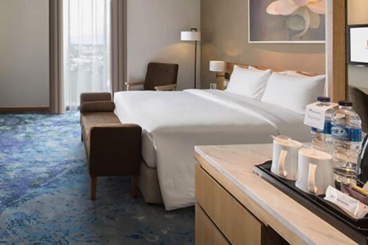 Ett hotellrum med en säng, ett skrivbord, en stol, ett litet bord med diverse bekvämligheter och ett stort fönster.