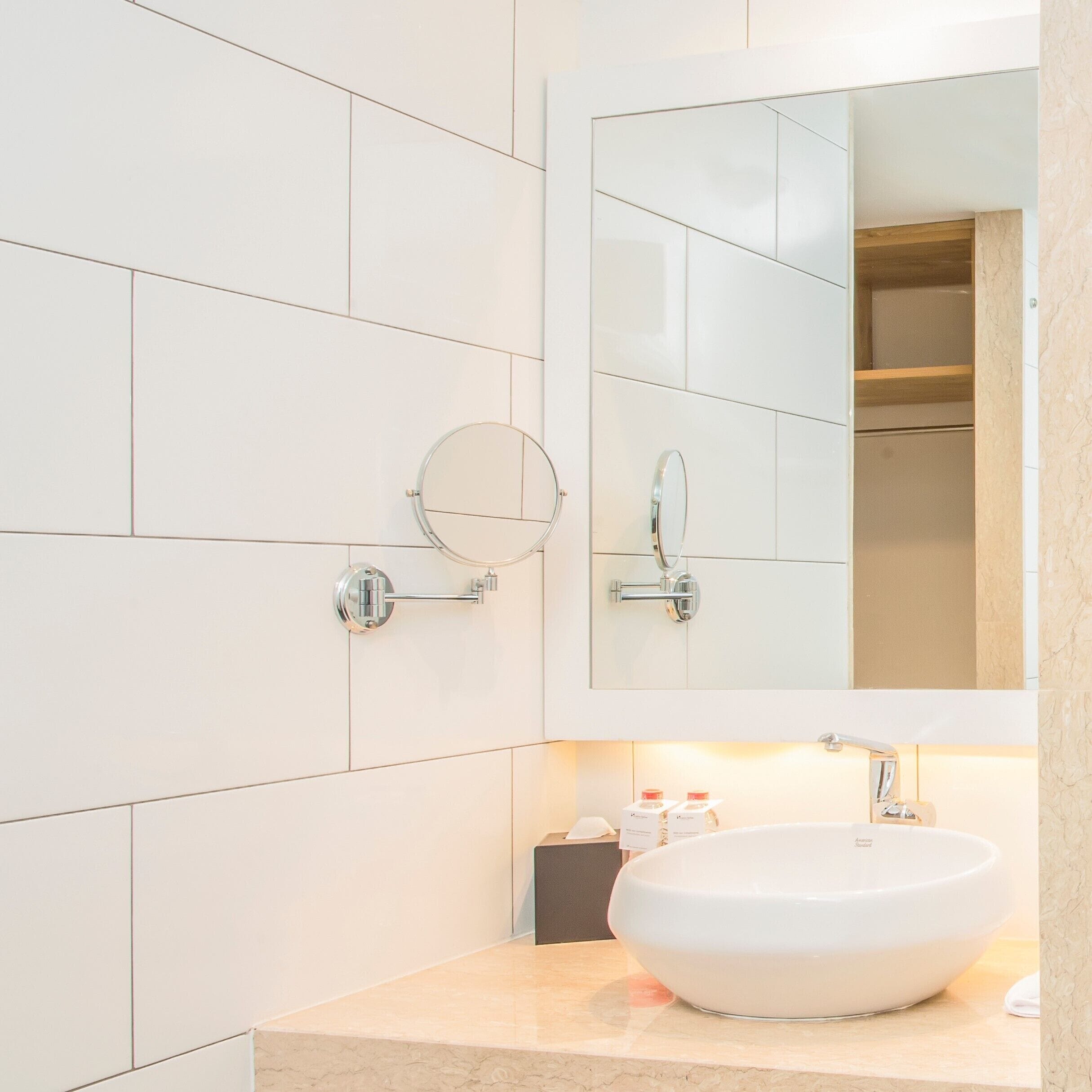 A modern bathroom with white tiles, a large mirror, a round mirror, a white sink, and a towel rack.