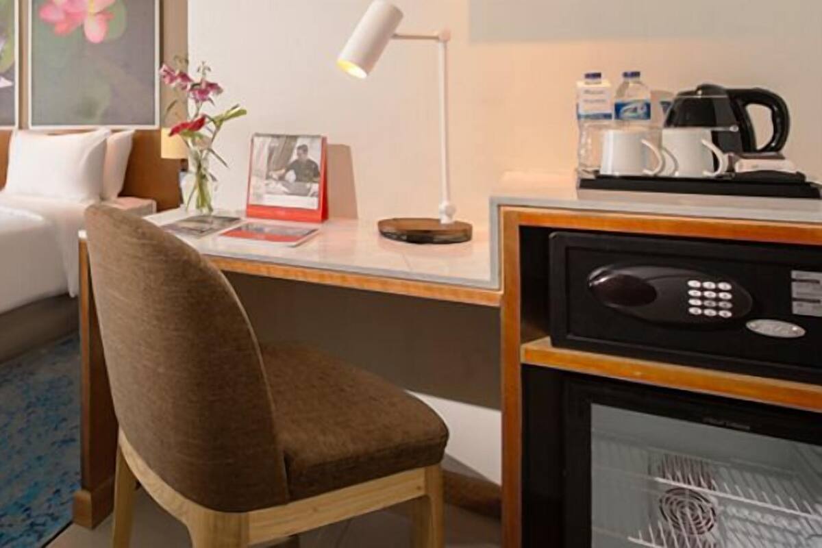 Ett hotellrum med en säng, ett skrivbord med en lampa, en stol, en kaffebryggare och ett kylskåp.
