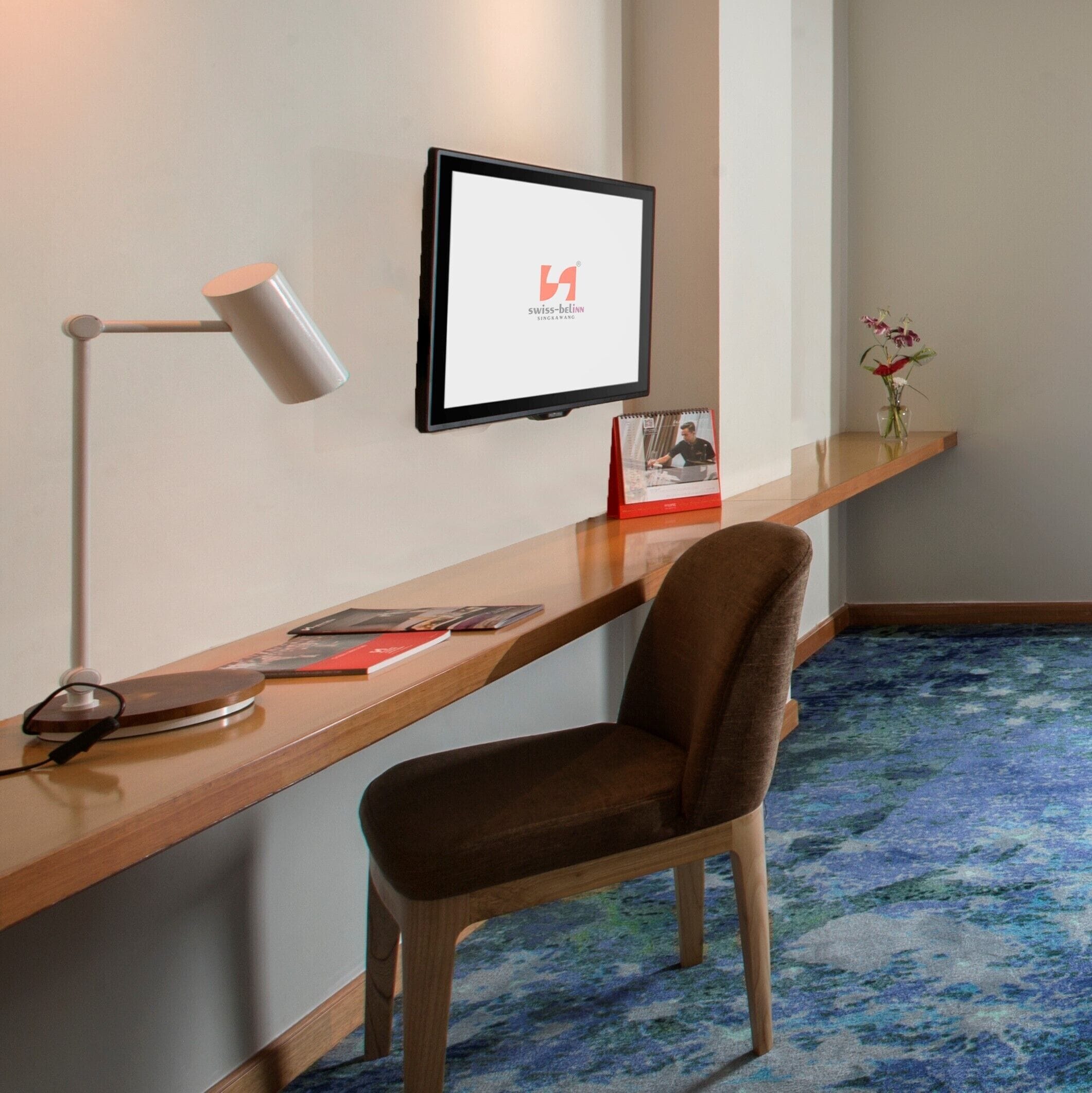 A hotel room with a wooden desk, a chair, a desk lamp, a framed picture, a small vase with flowers, and a carpeted floor.