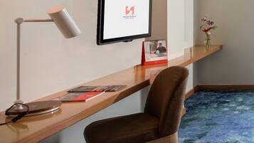 A hotel room with a wooden desk, a chair, a desk lamp, a framed picture, a small vase with flowers, and a carpeted floor.