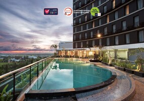 Una piscina infinita en la azotea, con barandilla de cristal, rodeada de exuberante vegetación y vistas a un paisaje urbano al atardecer.