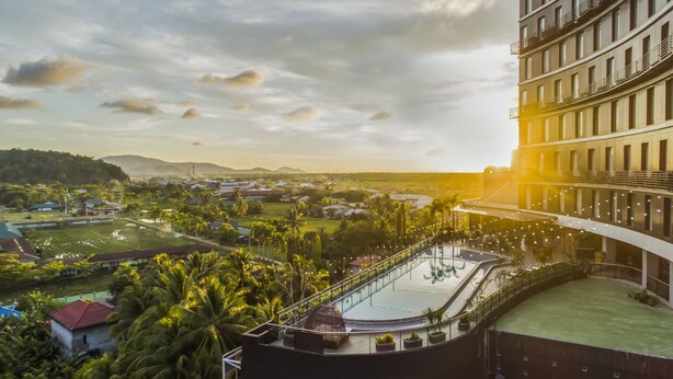 Un hotel moderno con piscina en la azotea, rodeado de vegetación exuberante y vistas a un pueblo distante.
