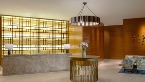 Un área de recepción con un mostrador de mármol, una pared decorativa con paneles iluminados, una lámpara colgante y sillas con estampado.