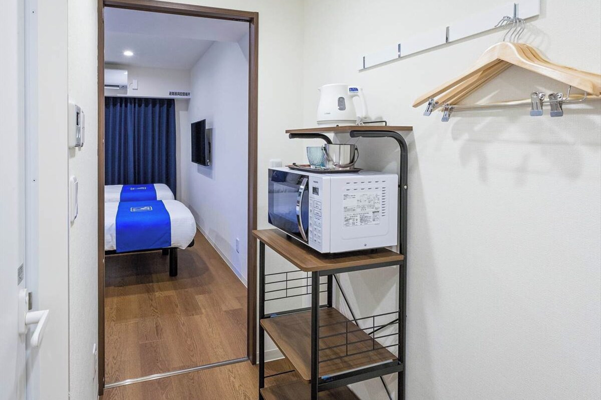 Habitación de hotel con cama, microondas, cafetera y repisas con perchas.