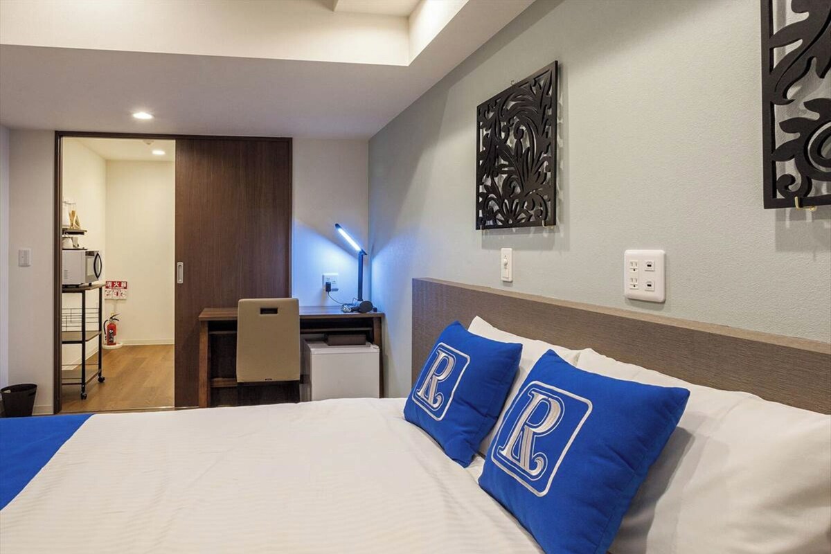Habitación de hotel con cama, escritorio y silla. Hay un adorno en la pared sobre la cama y un extintor en la pared.