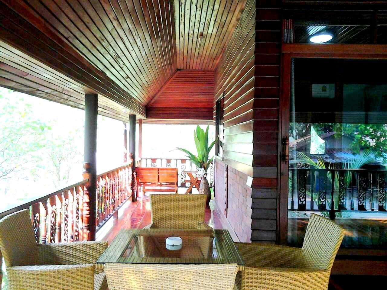 Un área de estar al aire libre techada con mobiliario de mimbre y una mesa de vidrio, con vistas a un jardín y un edificio.