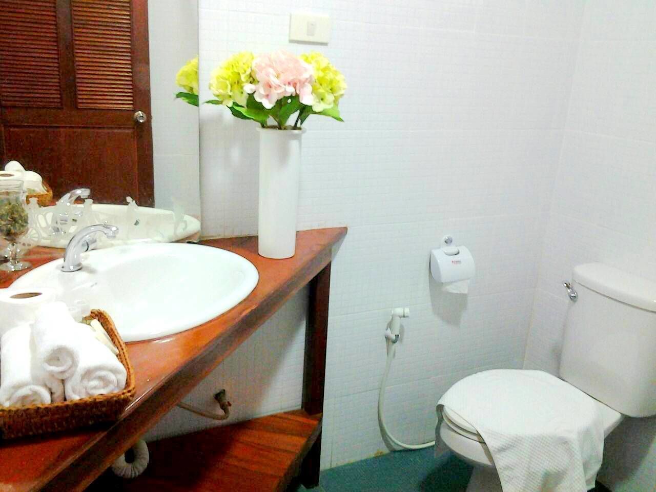 A bathroom with a wooden vanity, white sink, and a toilet. There is a vase with flowers on the vanity and a basket of towels.