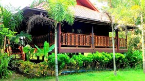 A traditional wooden house with a thatched roof, surrounded by lush greenery and a wooden deck.