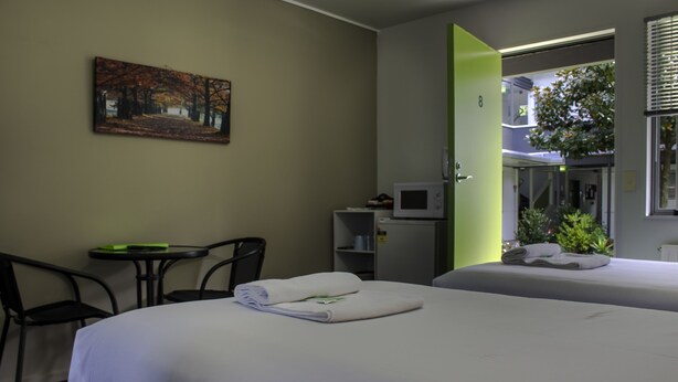 A hotel room with two beds, a desk, a chair, a painting on the wall, and a view of greenery through a window. - Te Henui Lodge (New Plymouth)