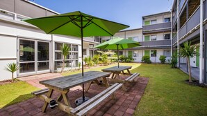 A courtyard with picnic tables and green umbrellas, surrounded by buildings with balconies and large windows. - Te Henui Lodge (New Plymouth)