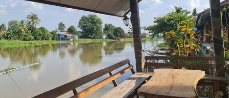 A wooden bench and table under a covered area overlooking a calm river with houses and greenery in the background.