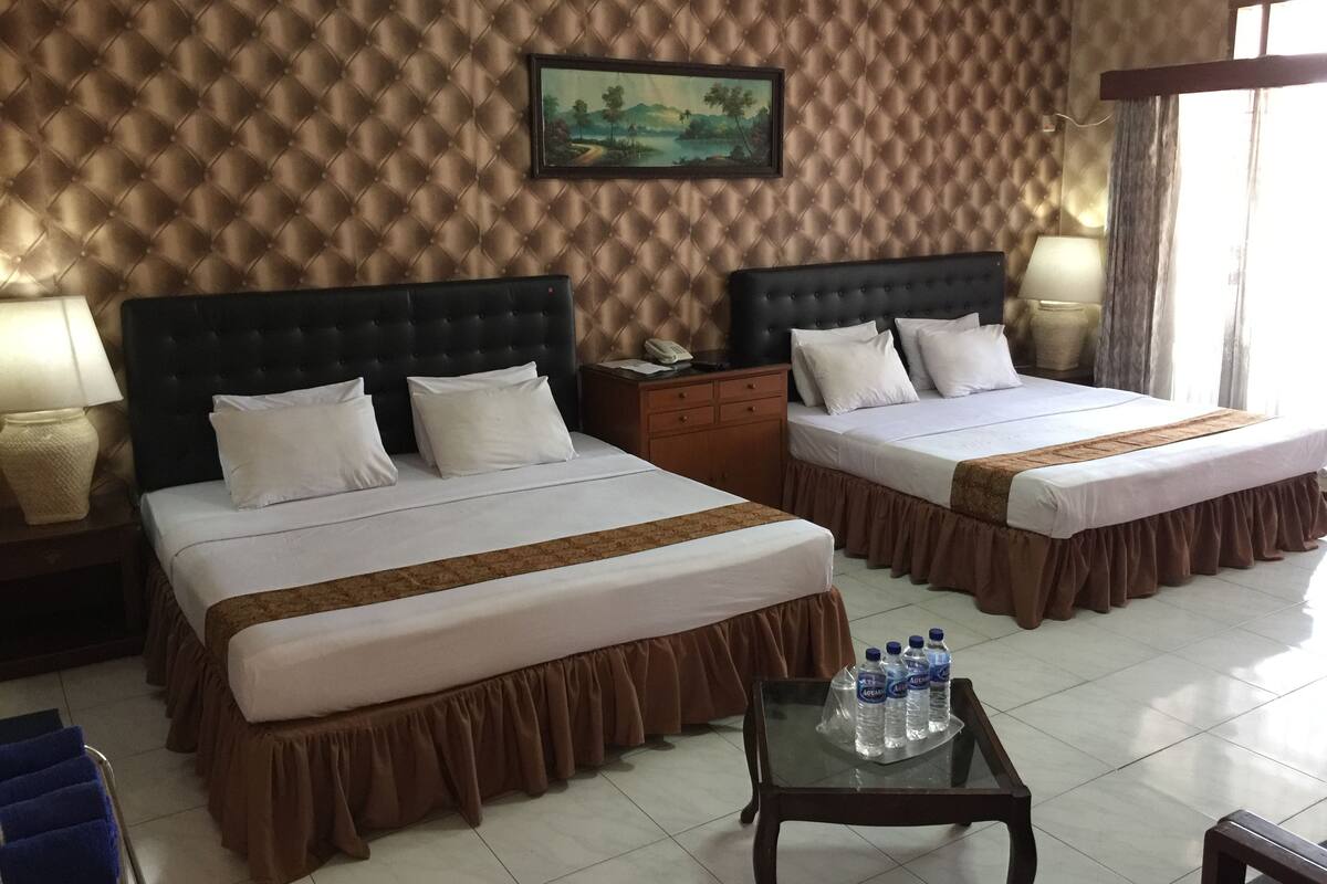 Habitación de hotel con dos camas, una mesita de noche, una lámpara, una mesa con botellas de agua y un cuadro en la pared.
