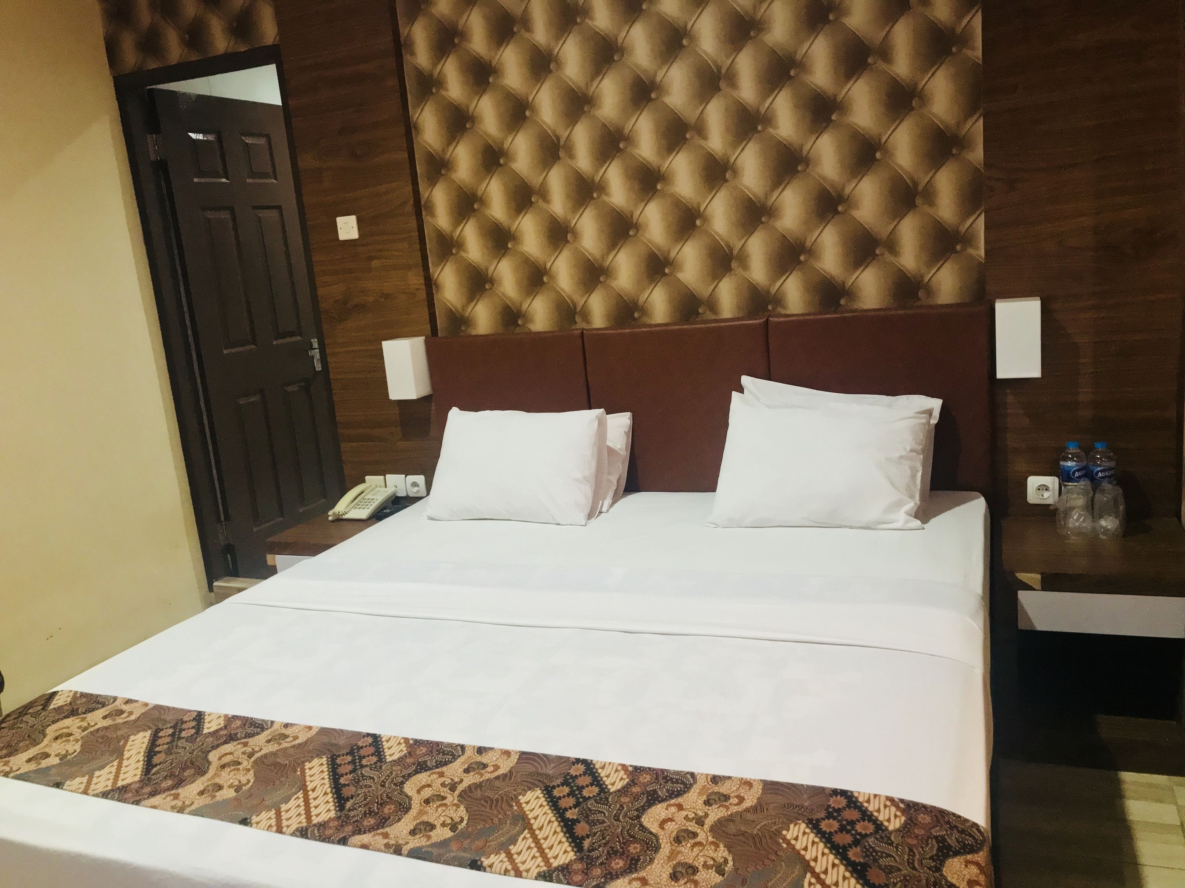 A hotel room with a large bed, a headboard with a tufted design, two bedside tables with lamps, a telephone, and two water bottles on the nightstand.