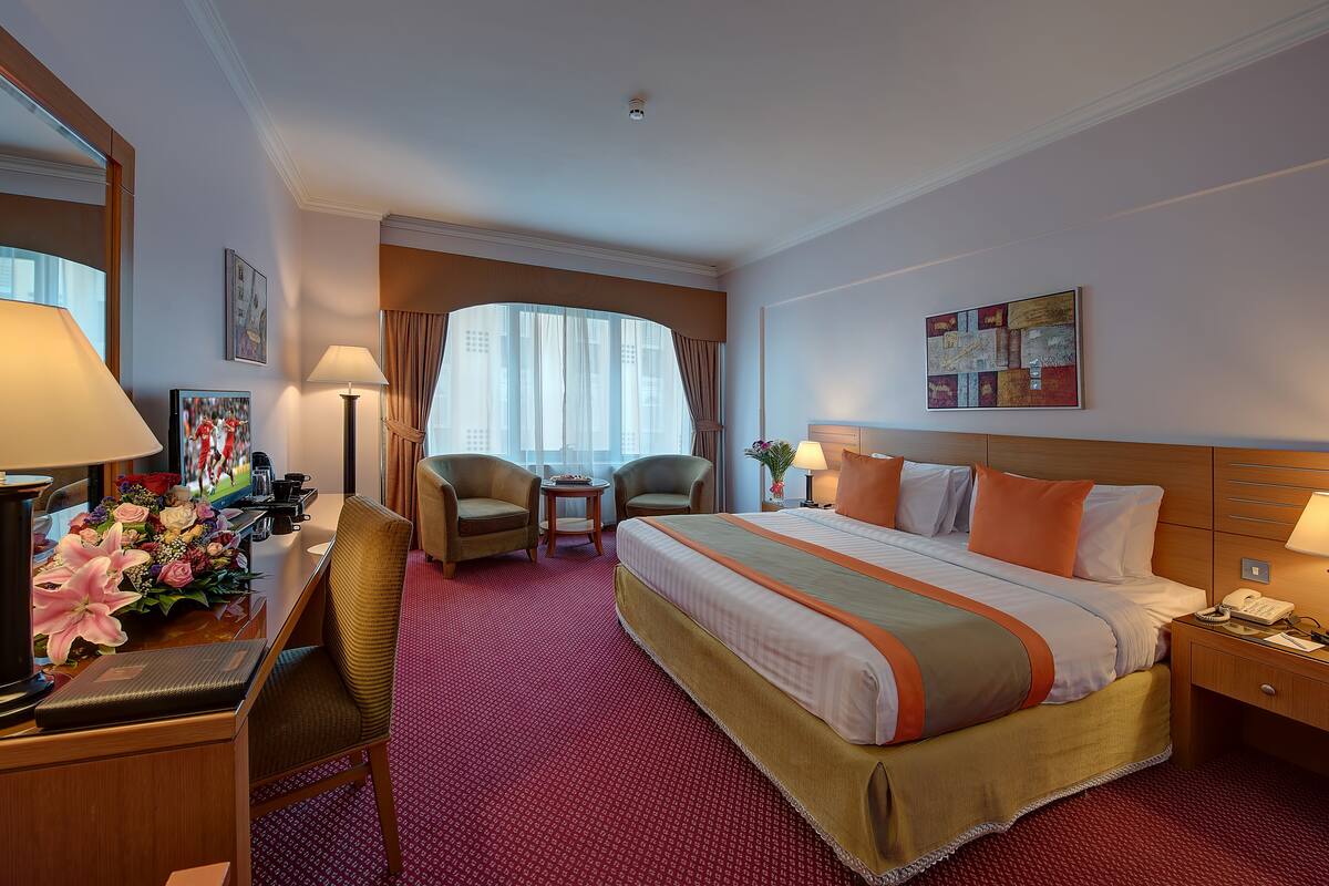 Une chambre d’hôtel avec un grand lit, deux fauteuils, un bureau avec une lampe et une vue sur la ville grâce à de grandes fenêtres.