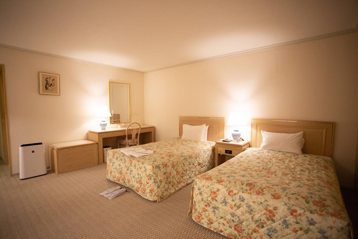 Una camera d'albergo con due letti, una scrivania, una sedia, un comodino, una lampada e un piccolo radiatore.