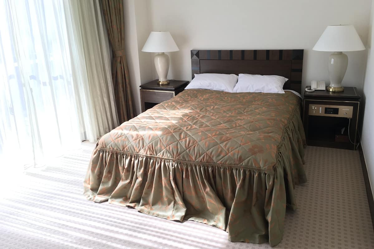 Una camera d'albergo con un letto grande, due comodini con lampade, una finestra con tende e un pavimento tappezzato.