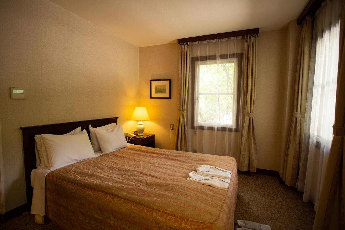 Una camera d'albergo con un letto grande, un comodino con una lampada, un quadro appeso al muro e una finestra con tende.