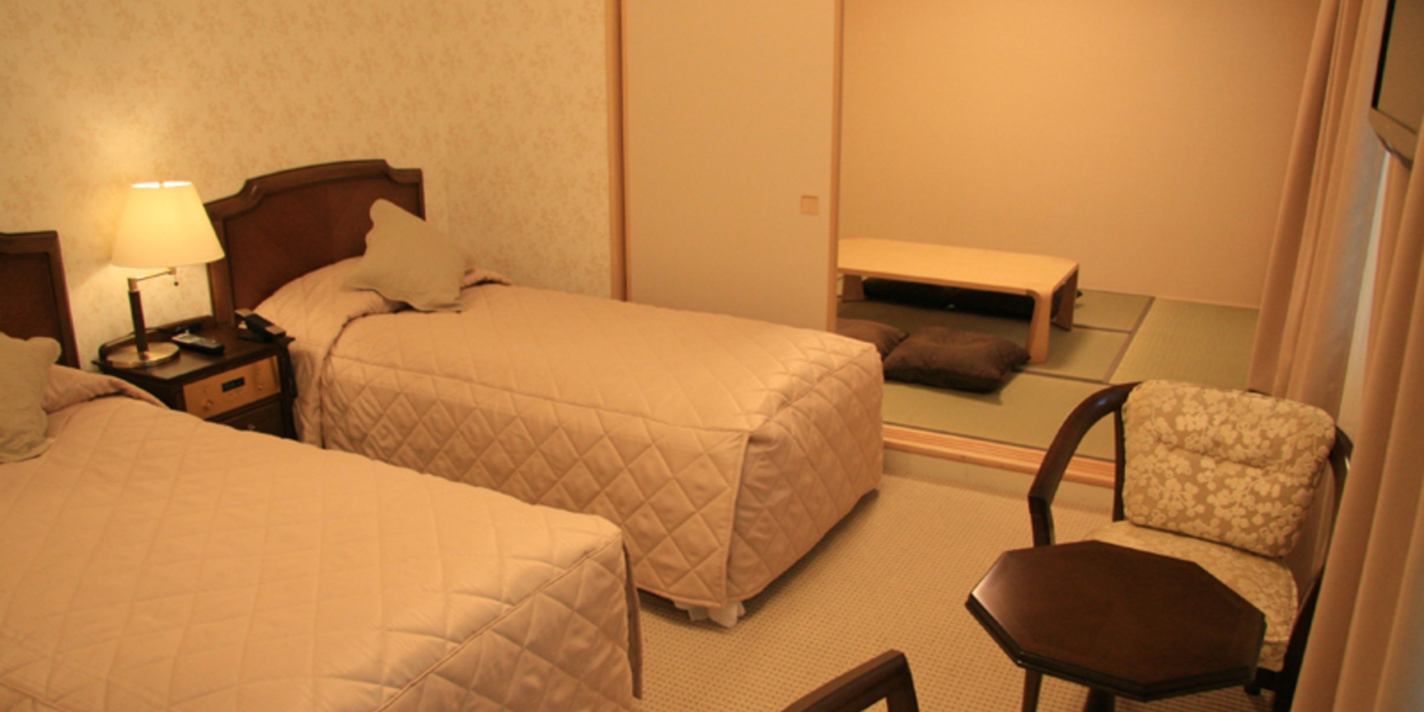 A hotel room with two beds, a nightstand with a lamp, a chair, and a small table.