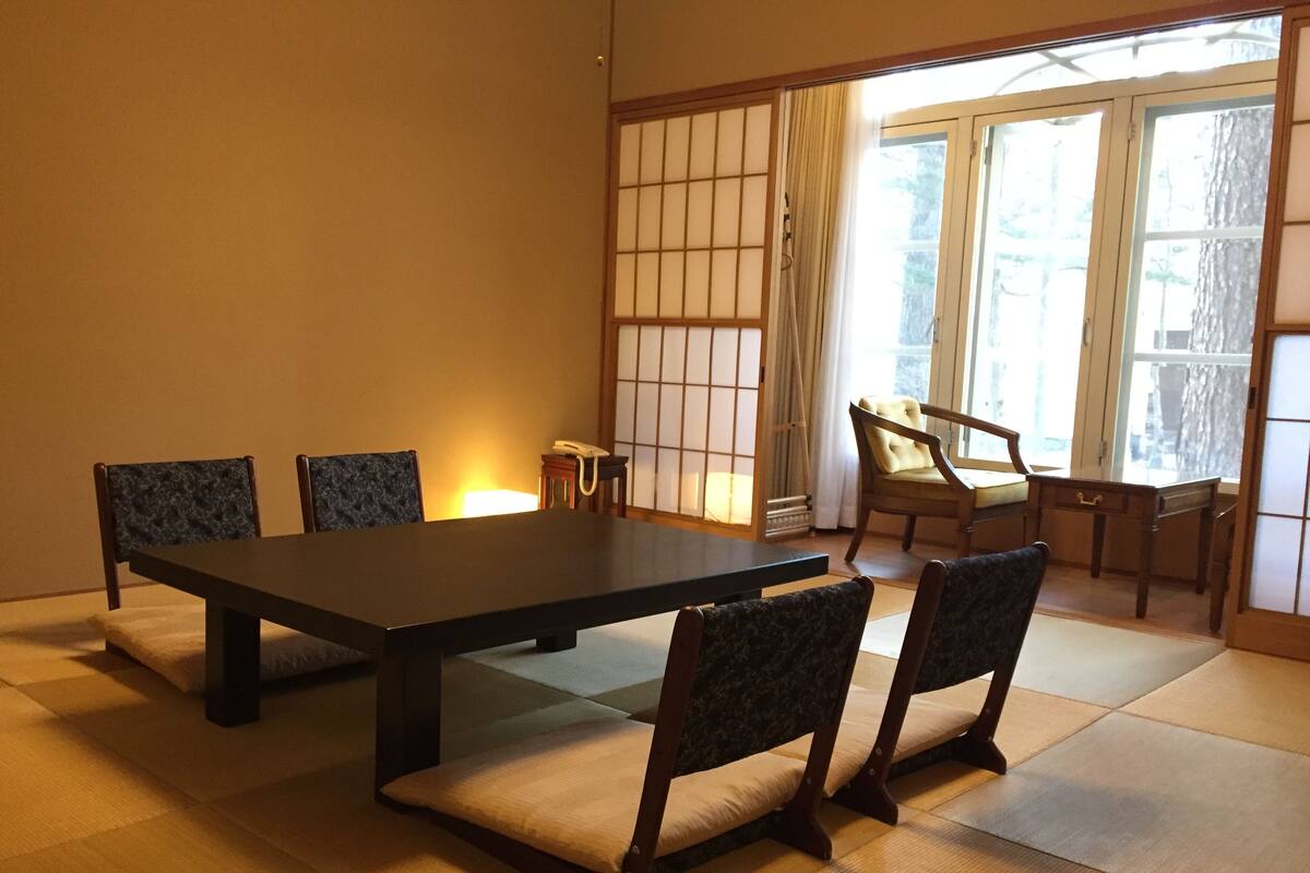 Una stanza tradizionale giapponese con pavimento in tatami, un tavolino e quattro sedie con cuscini.