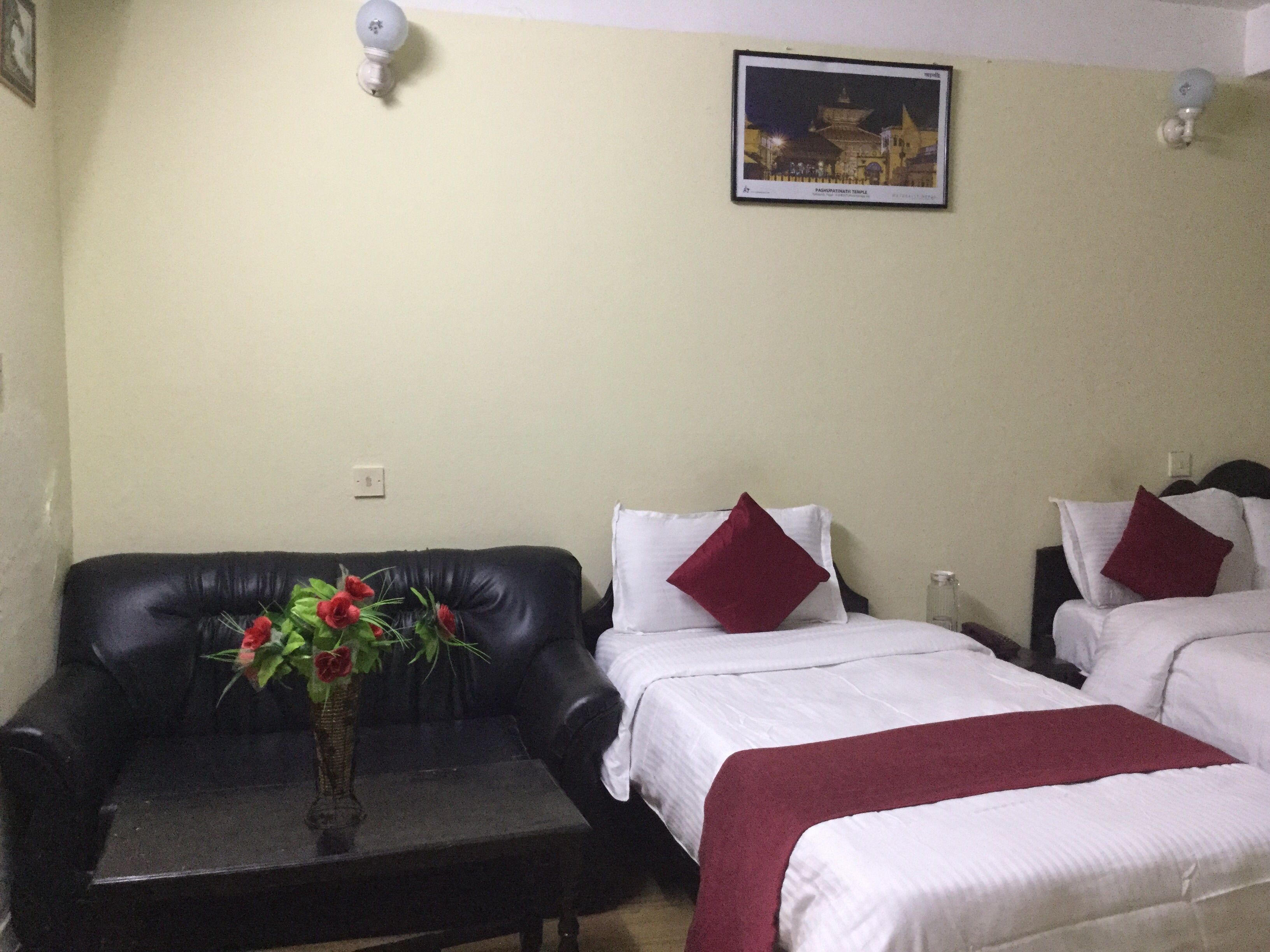 A hotel room with a black leather sofa, two beds with white bedding and red pillows, a small table with a vase of flowers, and a framed picture on the wall.