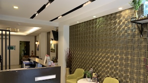 Zona de recepción con pared decorada, mostrador de recepción, dos sillas y una pequeña mesa con un florero y un marco de fotos.