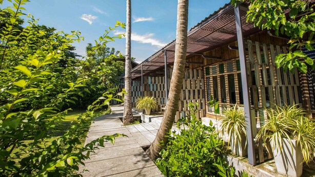 A pathway leading to a wooden building with a tiled roof, surrounded by lush greenery and tall palm trees.