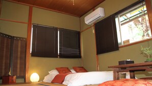 A traditional Japanese-style room with tatami flooring, a bed with white bedding and two red pillows, a wooden desk, and a large window with blinds. - Wakayama Guest House Shido (Hashimoto)