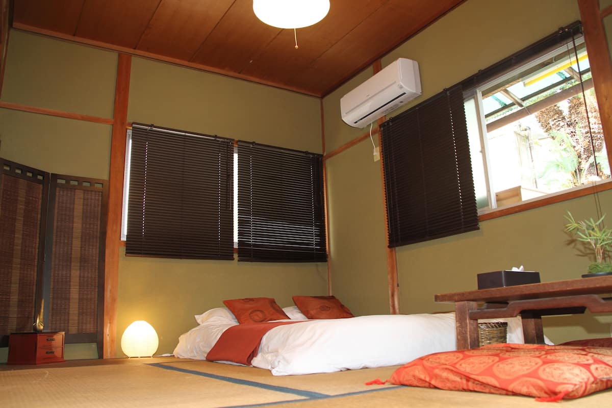A traditional Japanese-style room with tatami flooring, a bed with white bedding and two red pillows, a wooden desk, and a large window with blinds.