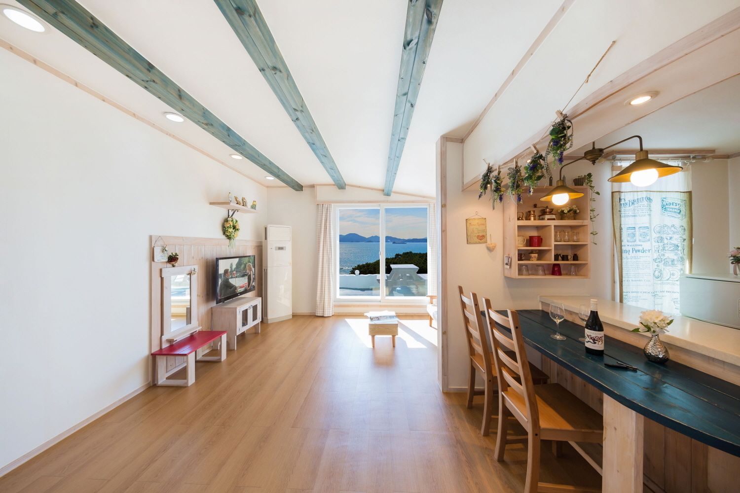Amplia sala de estar con suelo de madera, televisor, mesa de comedor con sillas y vista al mar a través de amplios ventanales.
