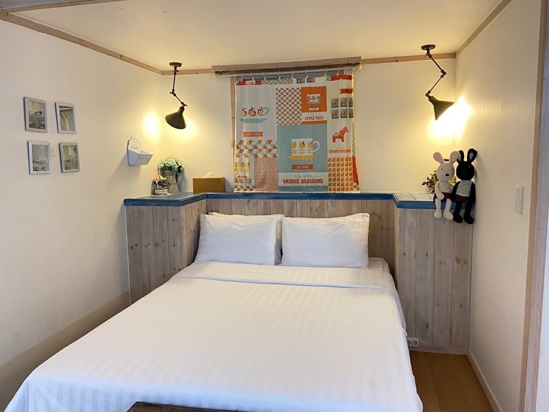 A bedroom with a wooden headboard, two bedside lamps, a quilted wall hanging, and a small shelf with decorative items.