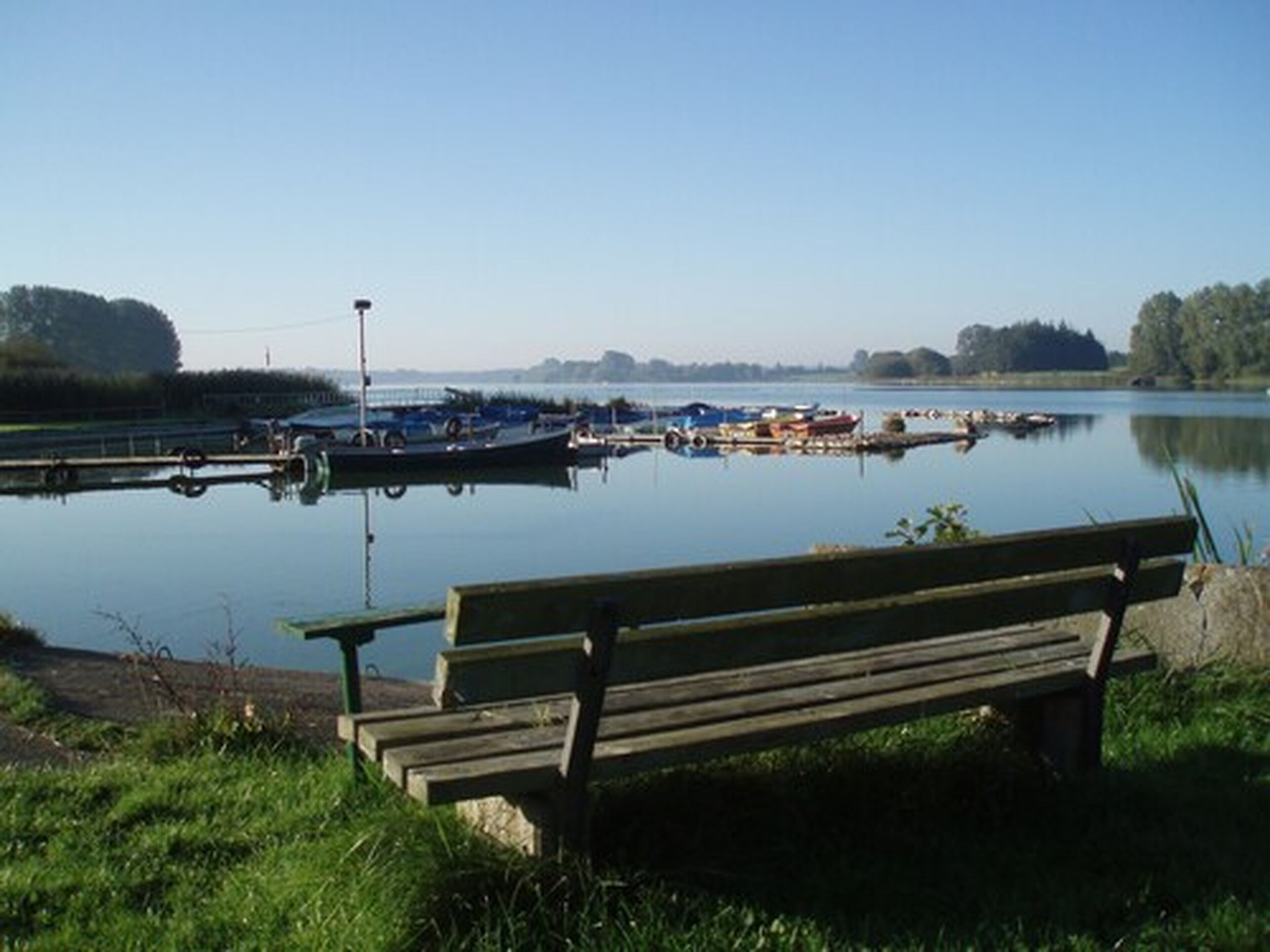 Ein ruhiger See mit vertäuten Booten, einer hölzernen Bank und blauem Himmel.