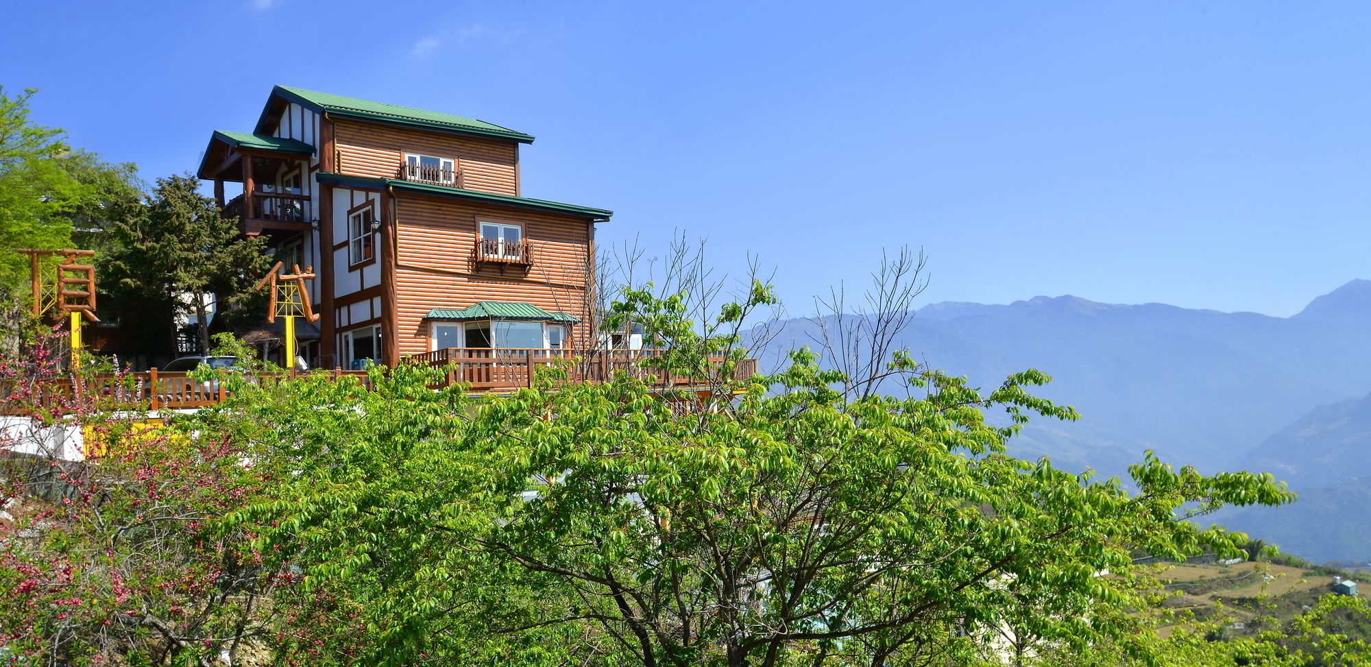 A two-story wooden building with balconies, surrounded by lush greenery and mountains in the background.