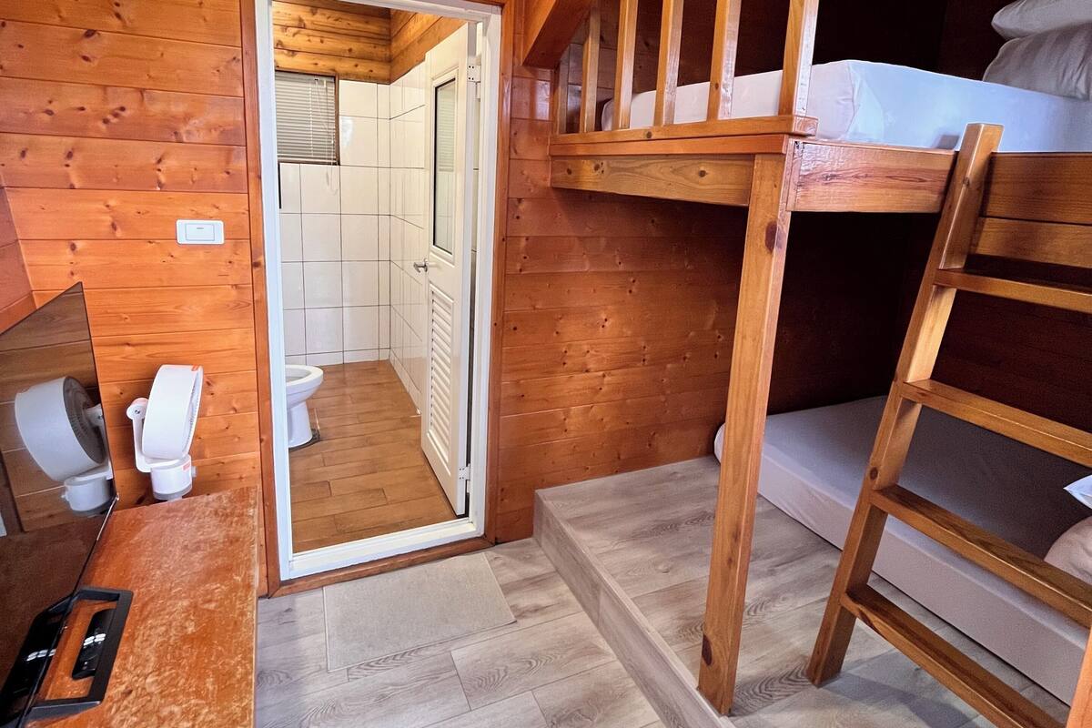 A wooden cabin interior with a bunk bed, a desk with a chair, and a bathroom visible through an open door.