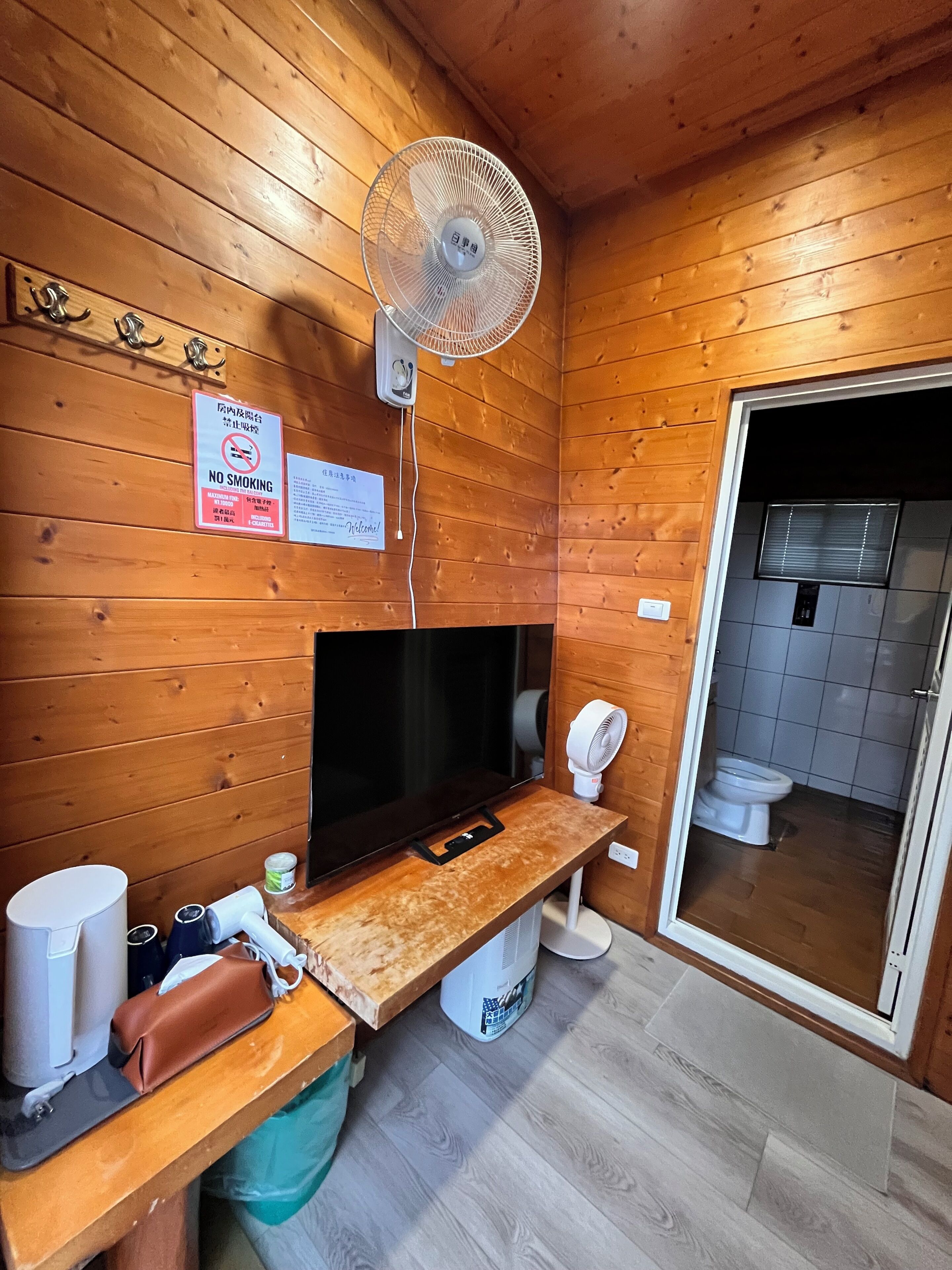 Habitación tipo cabaña de madera con televisión, ventilador, inodoro y un cartel que prohíbe fumar.