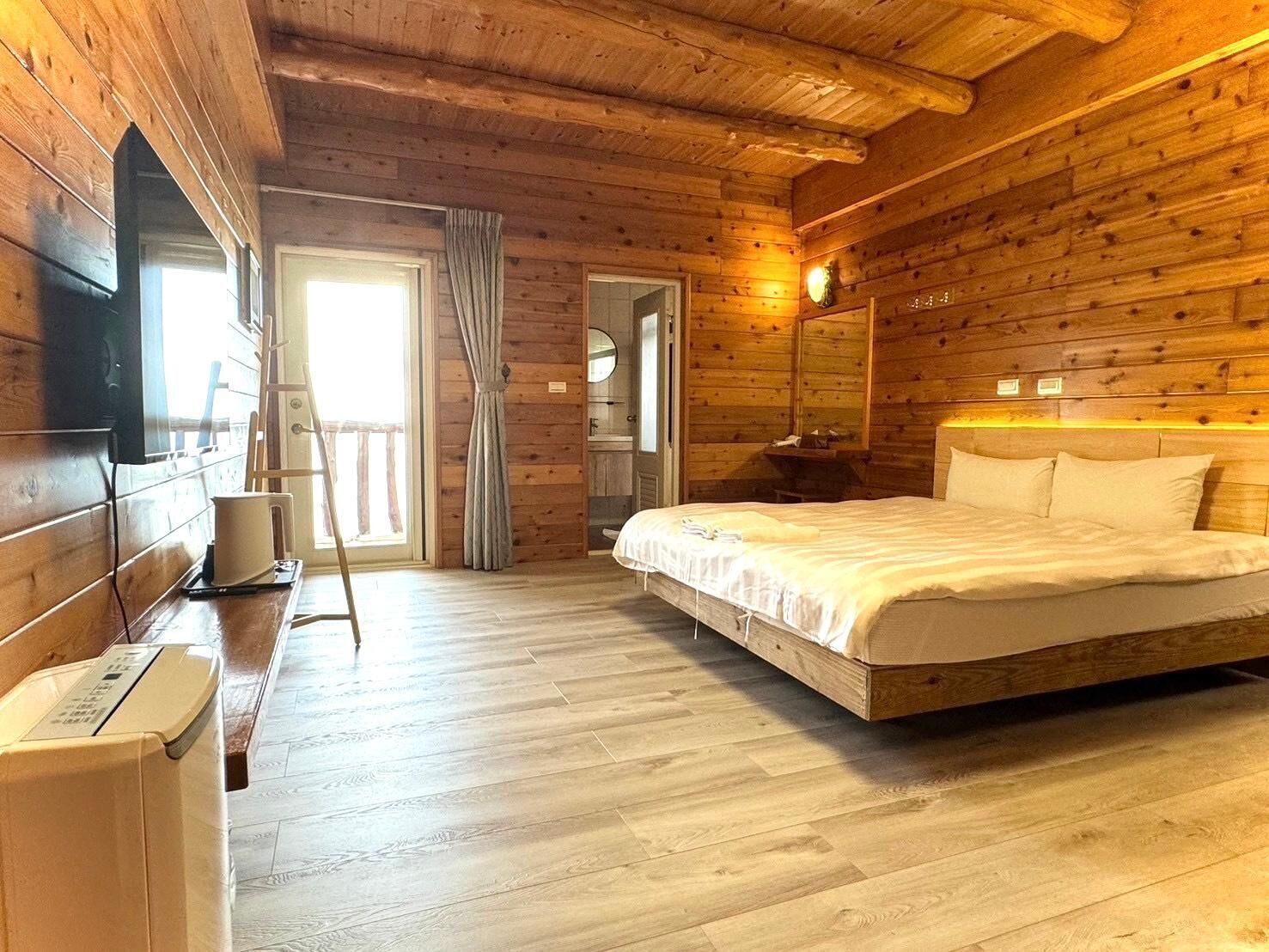 A wooden cabin room with a bed, a small table, a chair, a mirror, and a window.