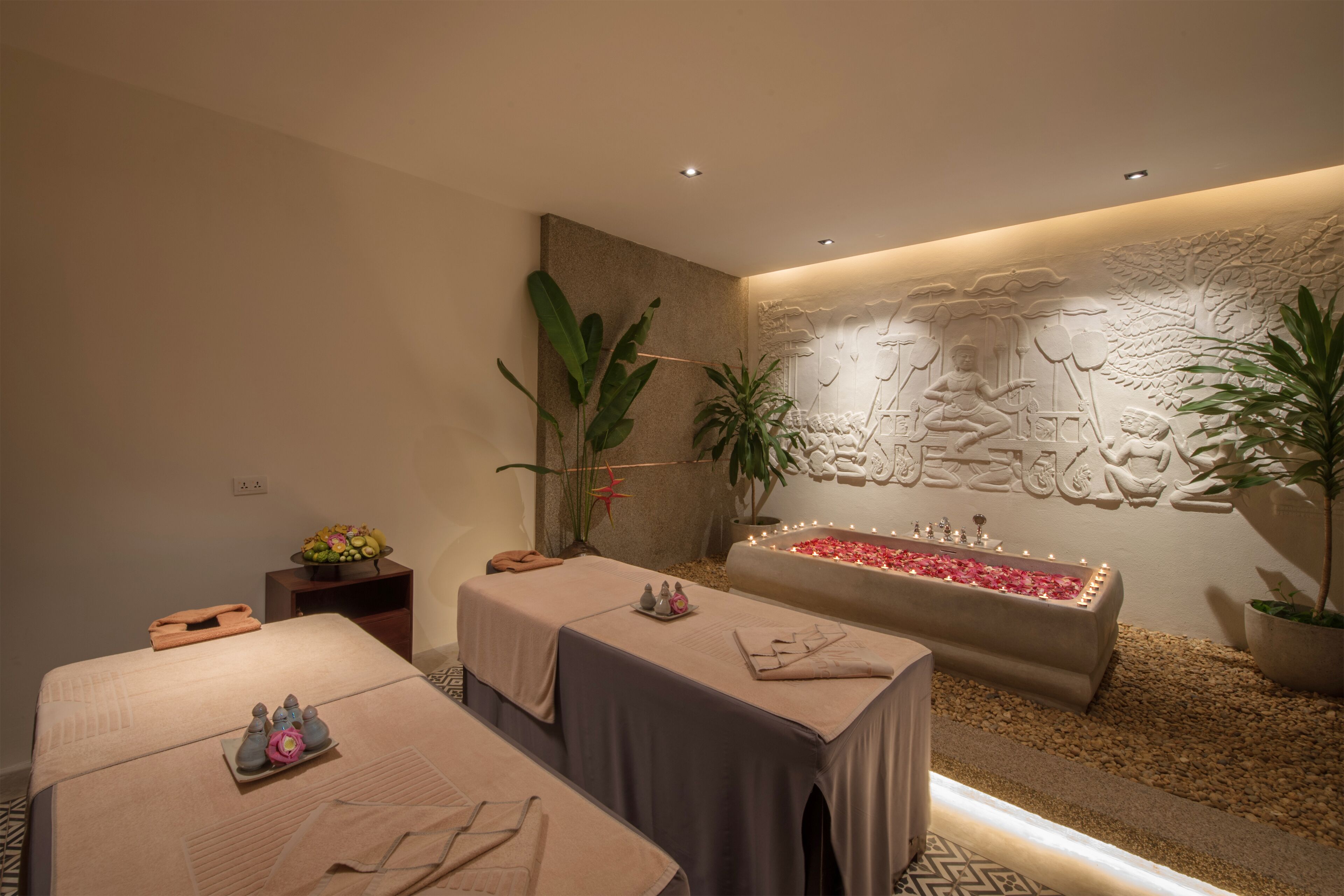 Un cuarto de spa con una camilla de masaje, una bañera con pétalos de rosa y una pared con un relieve esculpido.