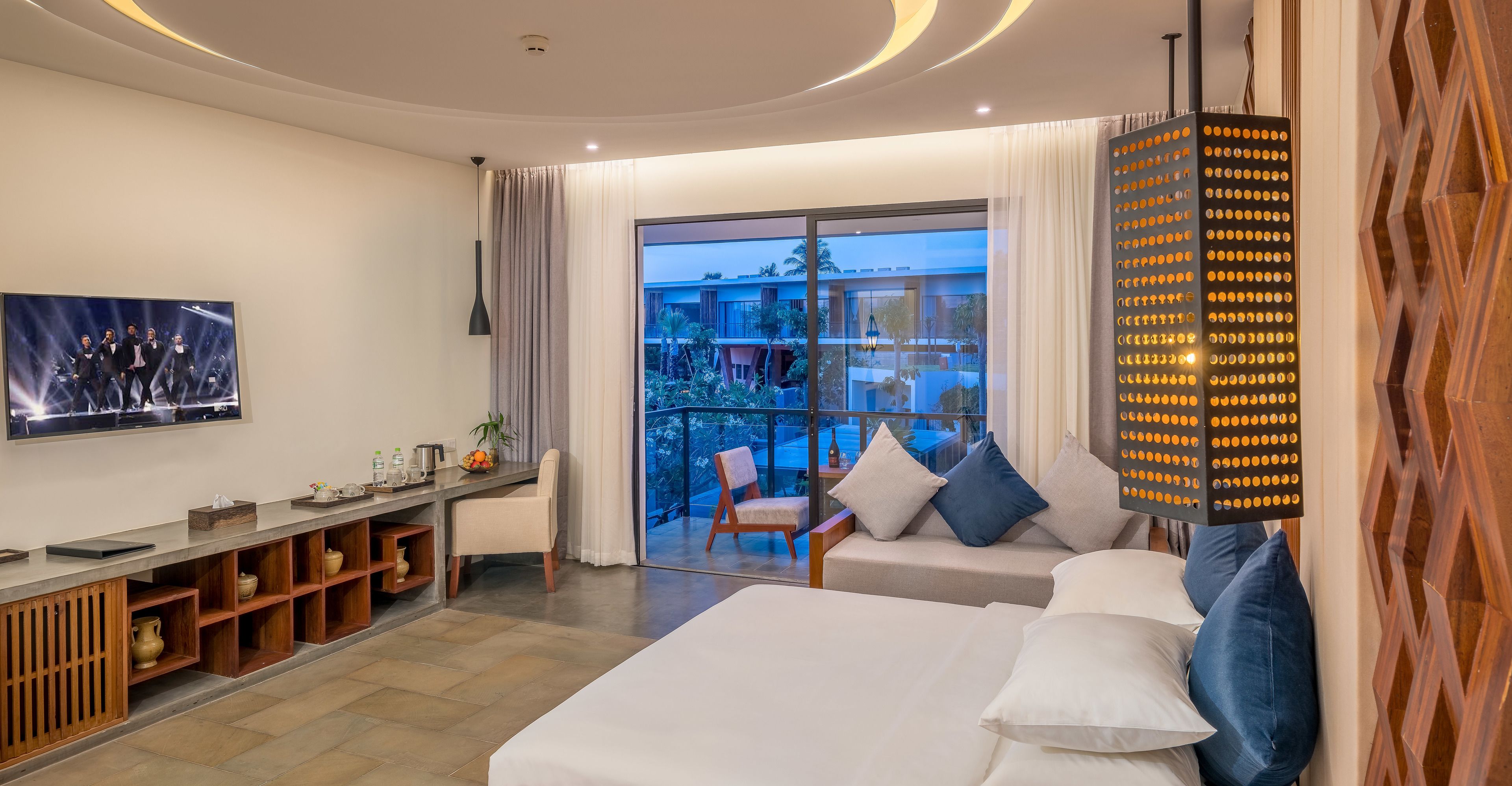 A modern hotel room with a large bed, a desk with a computer, a TV, and a balcony with a view of the city.