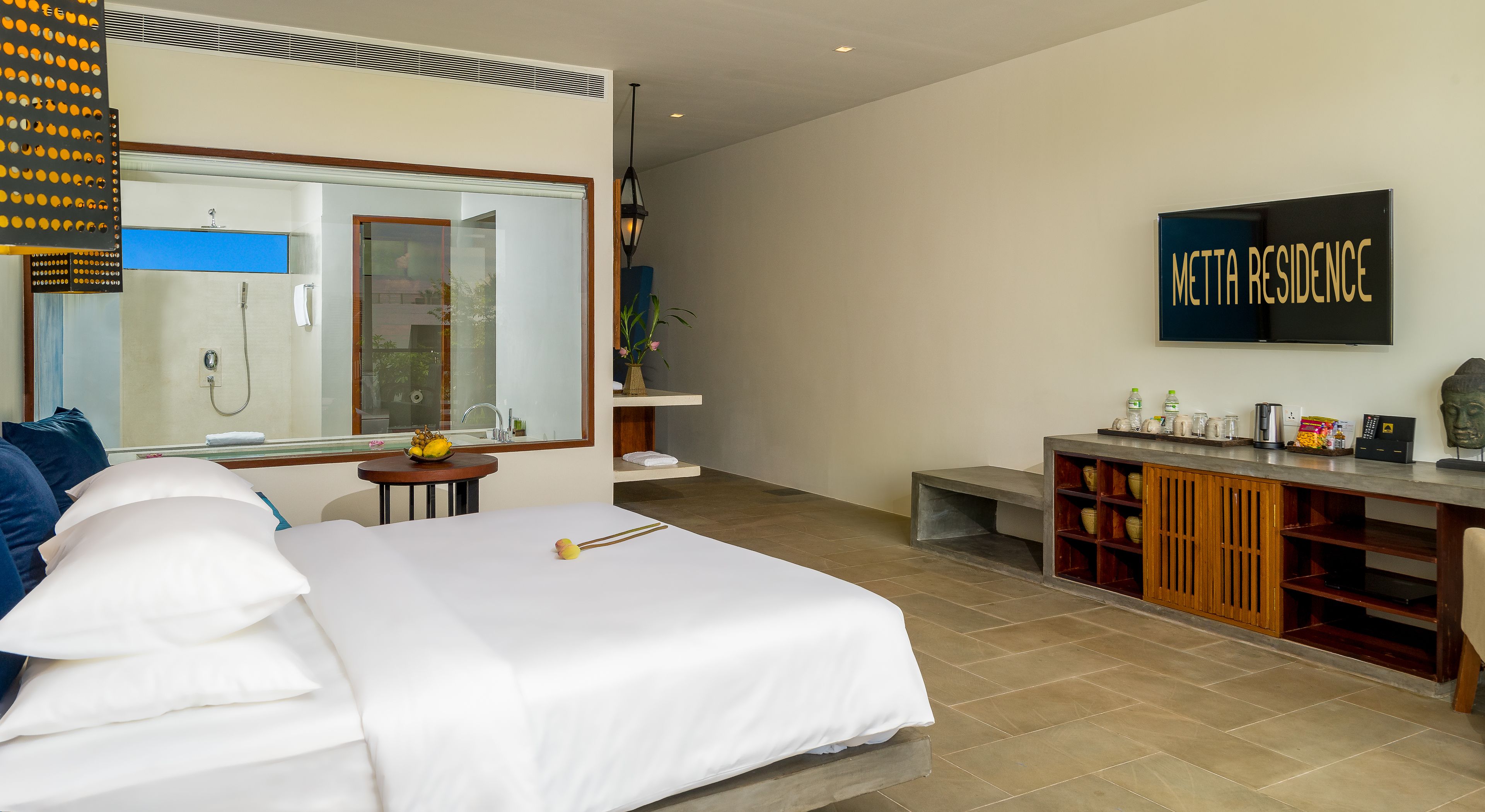 A modern hotel room with a large bed, a bathroom with a shower, a small table with a bowl of fruit, and a TV displaying "METTA RESIDENCE".
