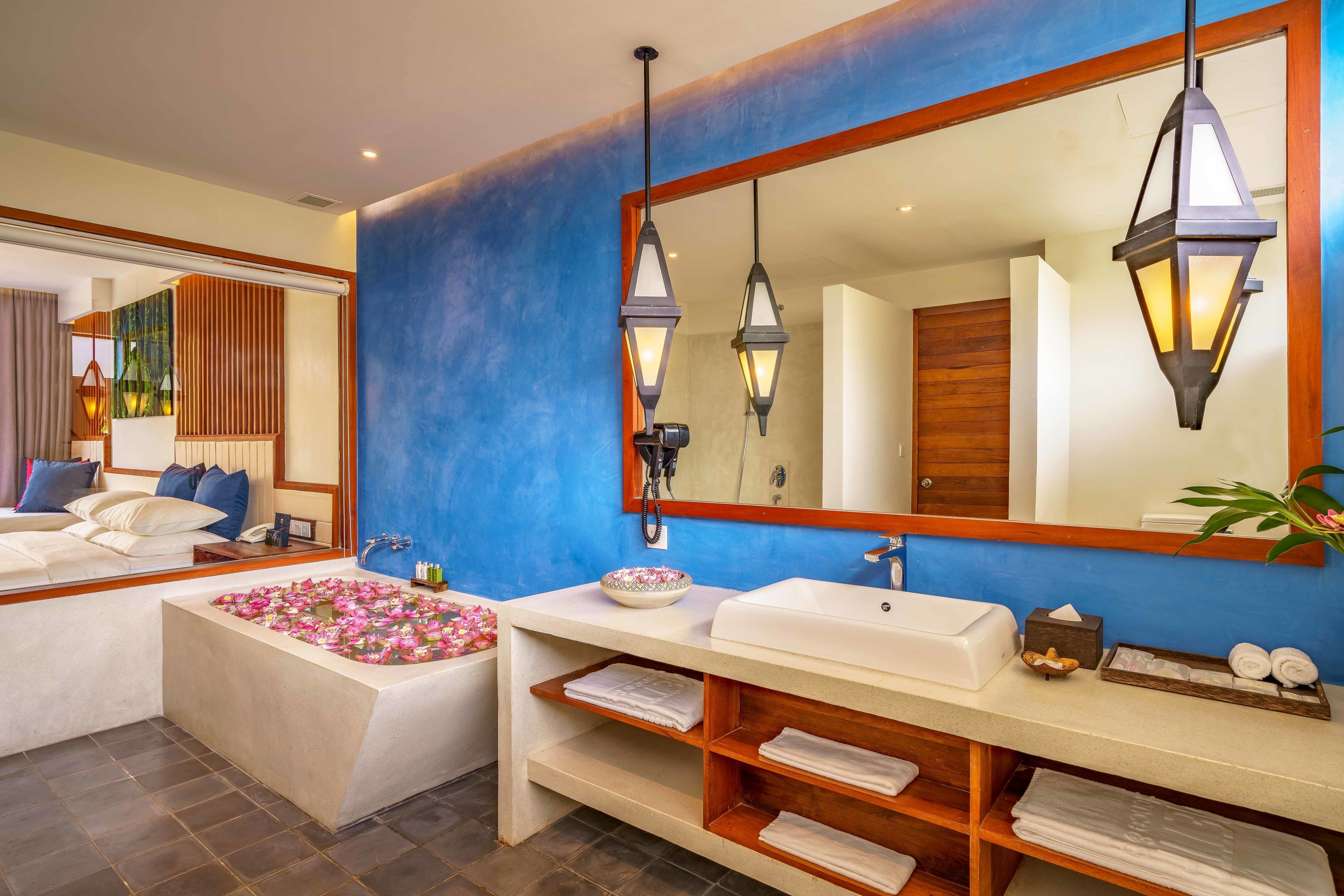 A modern bathroom with a large mirror, two pendant lights, a bathtub with flower petals, a white sink, and a wooden vanity.