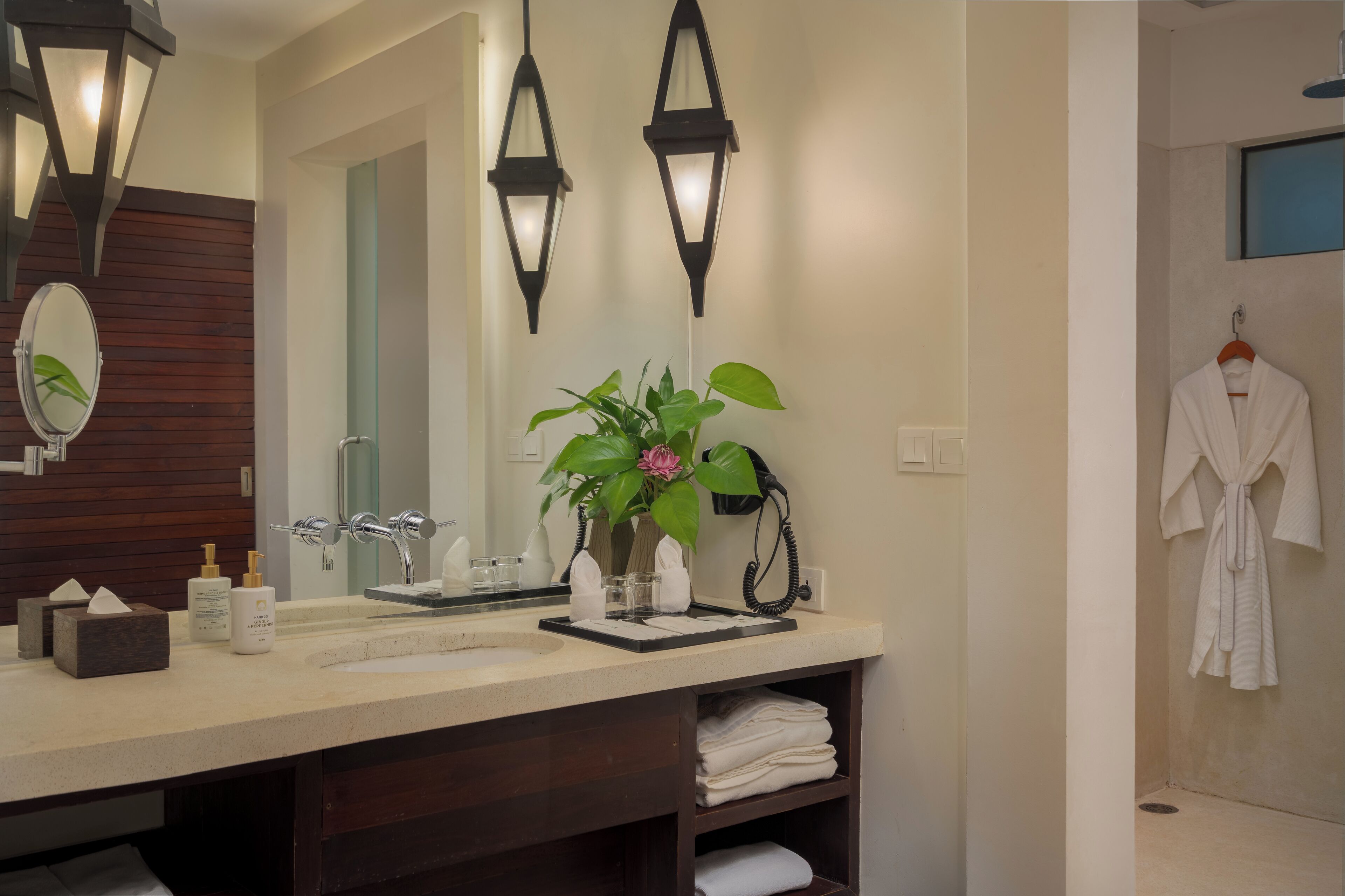 A modern bathroom with a large mirror, two wall-mounted lights, a sink with toiletries, a potted plant, and a robe hanging on the wall.
