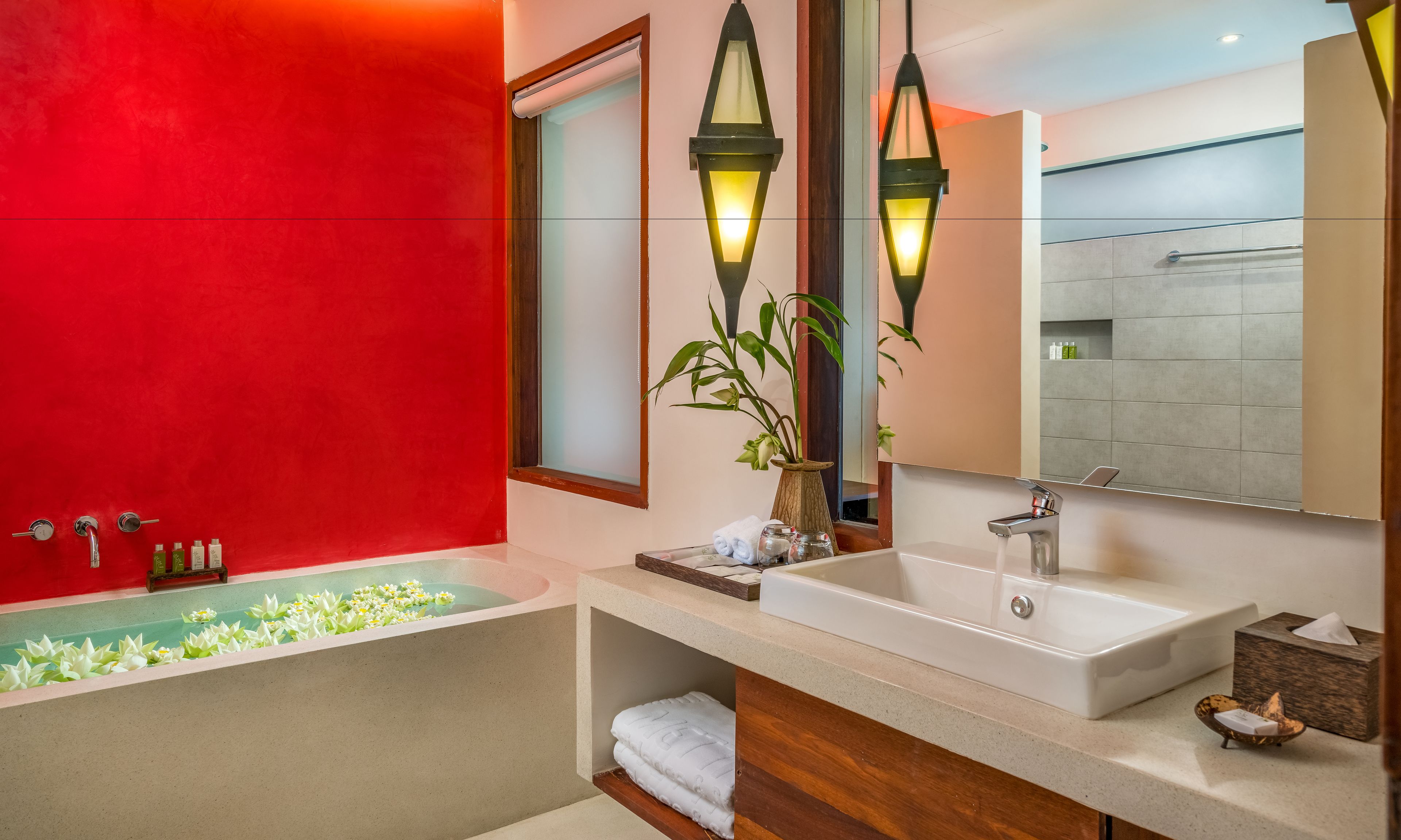 A modern bathroom with a red accent wall, a freestanding bathtub, a large mirror, and a wooden vanity.
